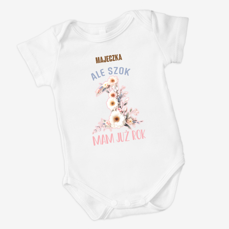 Biały body dla niemowląt z nadrukiem, na którym widnieją napisy: 'MAJECZKA', 'ALE SZOK', duża cyfra '1' ozdobiona kwiatami oraz 'MAM JUŻ ROK'.