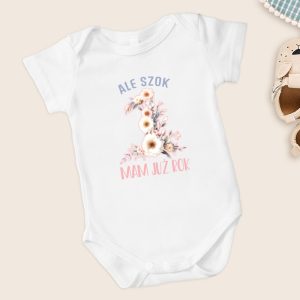 Biały body niemowlęce z napisem "ALE SZOK 1 MAM JUŻ ROK", ozdobione kwiatami, ustawione na jasnym tle, obok małych bucików.