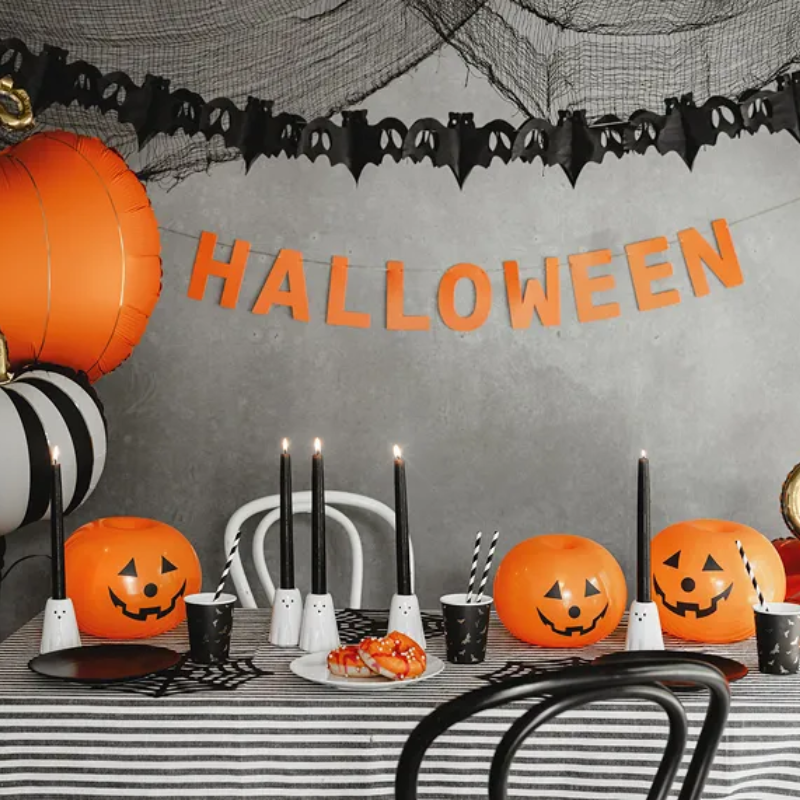 Stół udekorowany na Halloween, z czarnymi świecami, pomarańczowymi dyniami z wyciętymi twarzami, czarnymi kubkami w białe pająki oraz talerzem z przysmakami, w tle banner z napisem 'HALLOWEEN' i batwyką dekoracyjną.