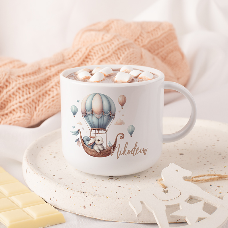 Biały kubek z rysunkiem balonu i małego misia, pełen gorącej czekolady z piankami, na talerzu w kolorze jasnym z delikatnymi plamkami, w tle widoczna beżowa dzianina i kawałki białej czekolady.