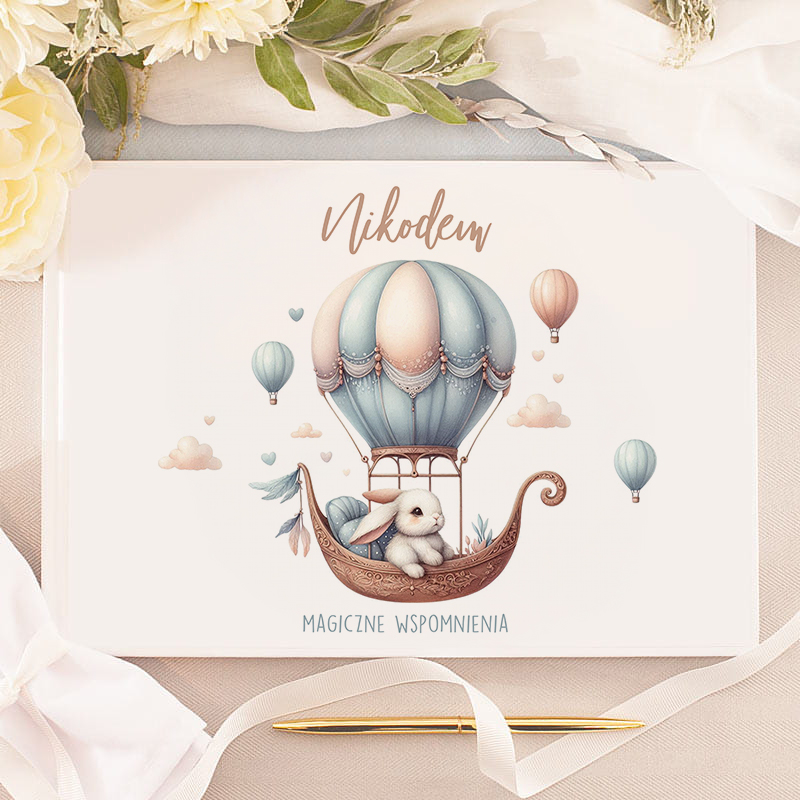 Ilustracja przedstawiająca króliczka siedzącego w koszu balonu na gorące powietrze, otoczonego balonami i chmurkami, z napisem 'Nikodem' i 'MAGICZNE WSPOMNIENIA' na białym tle.