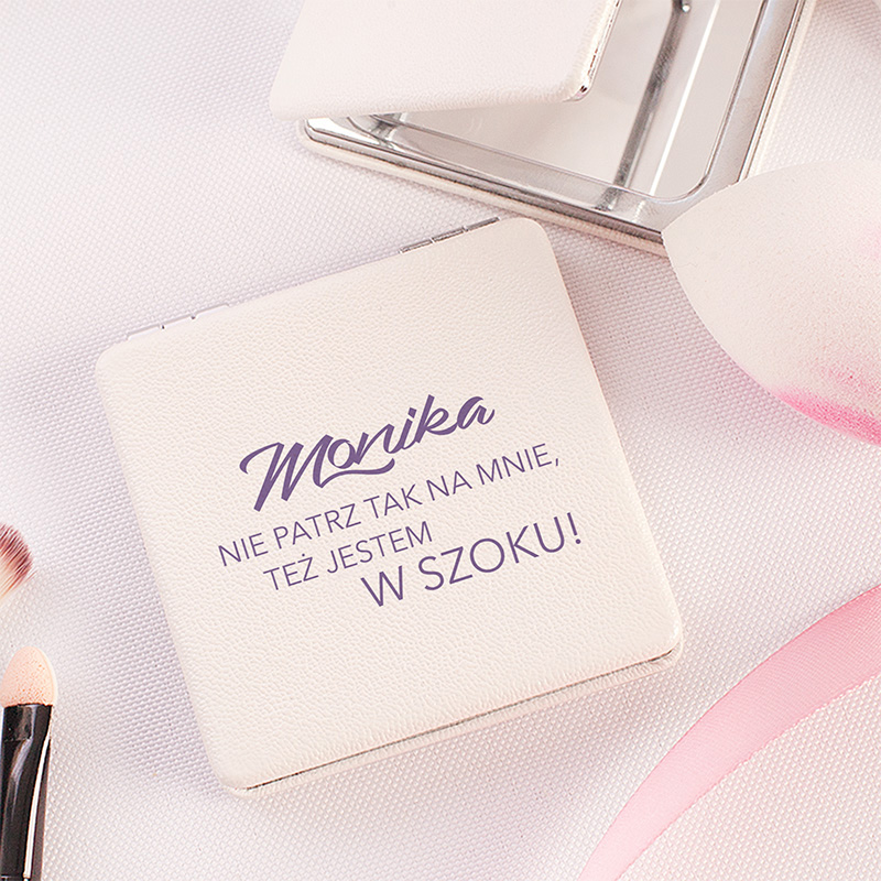 Lusterko w białym kolorze z napisem 'Monika' oraz tekstem 'NIE PATRZ TAK NA MNIE, TEŻ JESTEM W SZOKU!', obok pędzle do makijażu oraz gąbka do nakładania kosmetyków na jasnym tle.