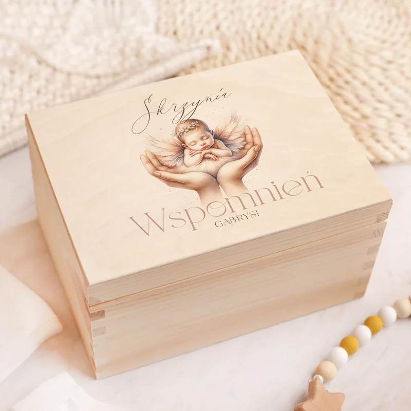 Drewniana skrzynia z napisem 'Skrzynia Wspomnień Gabrysi', na wieczku przedstawiająca ilustrację śpiącego dziecka z anielskimi skrzydłami w dłoniach opiekuna.