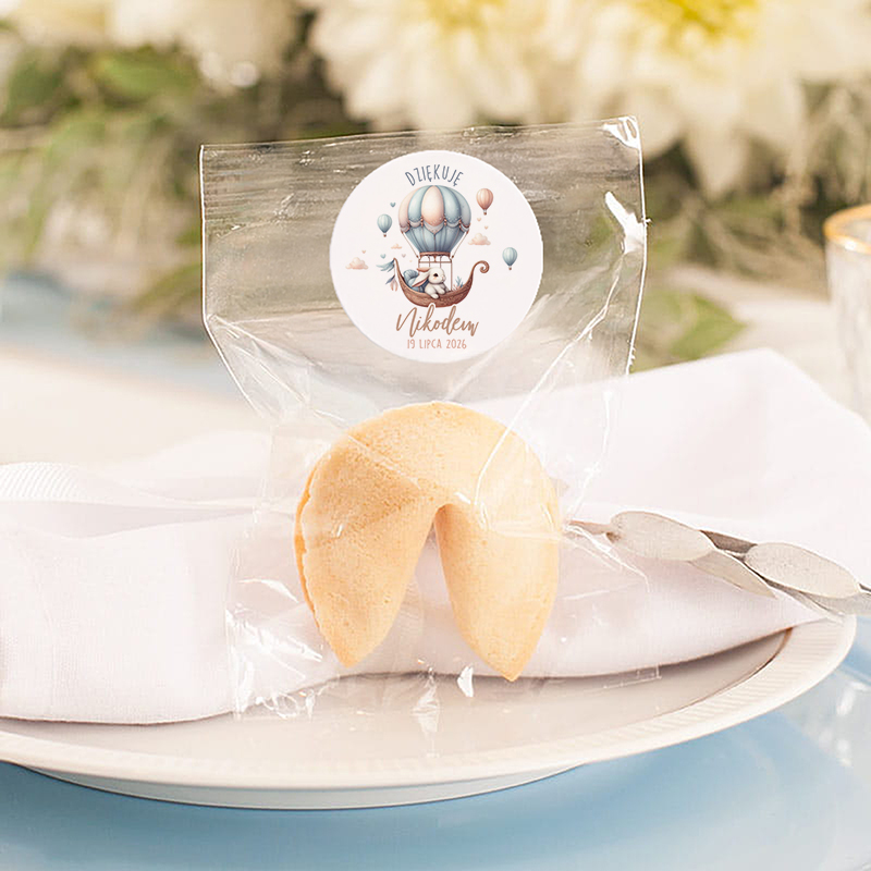 Ciasteczko szczęścia umieszczone w przezroczystym opakowaniu, z etykietą pokazującą ilustrację balonu na gorące powietrze oraz napisem 'Dziękuję Nikodem 19 lipca 2026'. Ciasteczko leży na białym serwetce wyłożonym na talerzu.