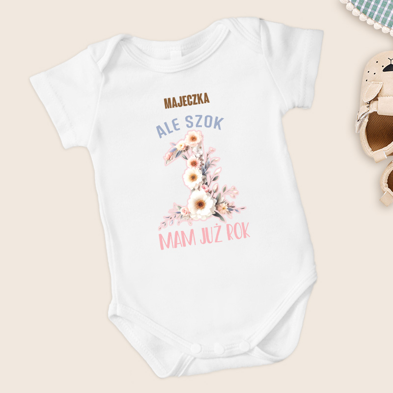 Biały body dziecięce z napisami "MAJECZKA ALE SZOK" i "MAM JUŻ ROK" w kolorze różowym i niebieskim, z cyfrą "1" ozdobioną kwiatami, obok pary dziecięcych bucików.