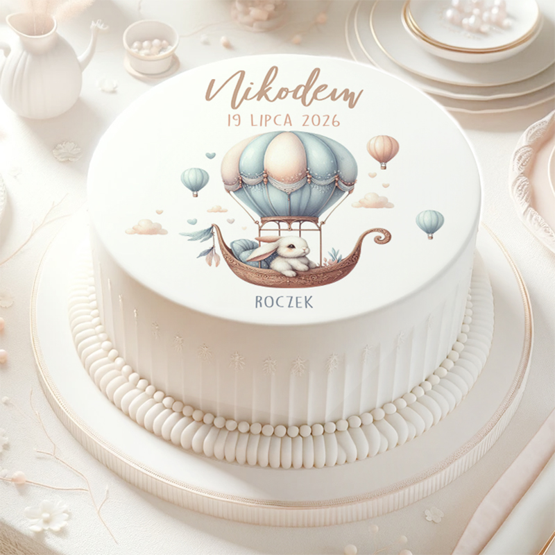 Elegancki tort z białą polewą, na którym znajduje się obrazek z balonem na gorące powietrze oraz króliczkiem. Na górze tortu napis "Nikodem 19 Lipca 2026" oraz słowo "Roczek". Tort jest ozdobiony delikatnymi, białymi detalami na boku.