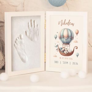 Ramka z odciskami dłoni i stóp dziecka, po lewej stronie widać odcisk dłoni i stopy w białym materiale, a po prawej stronie kolorowy projekt przedstawiający balon na gorące powietrze z króliczkiem i danymi urodzinowymi, takimi jak imię Nikodem, data urodzenia oraz waga i wzrost.