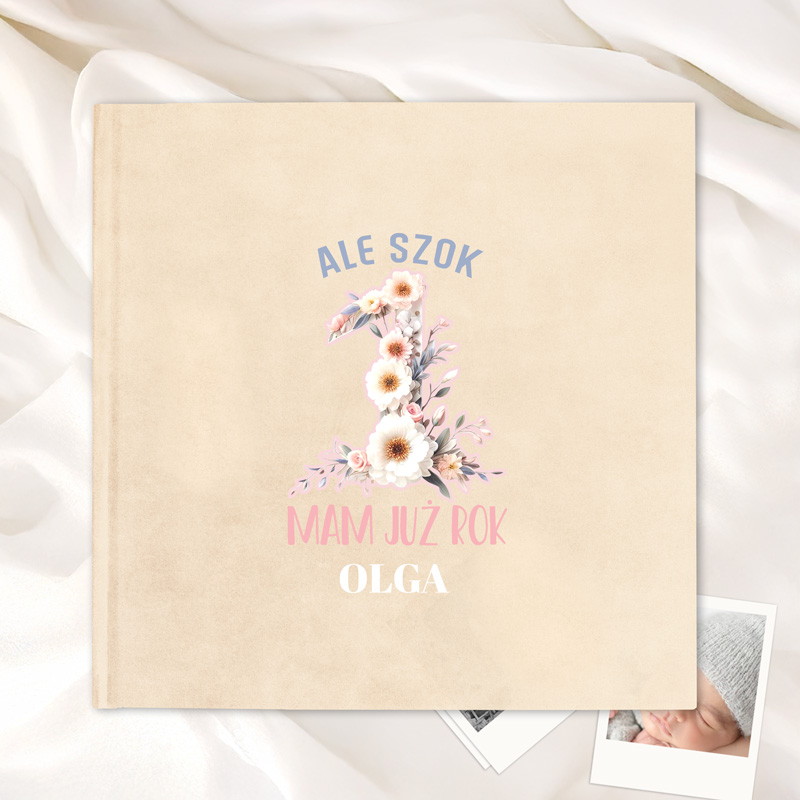 Okładka książki z napisem 'ALE SZOK' na górze, dużą liczbą '1' ozdobioną kwiatami w środku, tekstem 'MAM JUŻ ROK' poniżej i imieniem 'OLGA' na dole, umieszczona na jasnym tle z plisowanym materiałem obok.