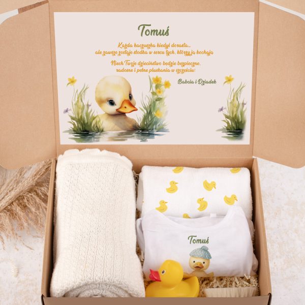 Pudełko z prezentem dla dziecka zawierające miękkie tekstylia oraz gadżety z motywem kaczki, z nadrukowanym imieniem "Tomuś". Wewnątrz widoczny jest list z rysunkiem kaczki i kwiatami na tle wody.