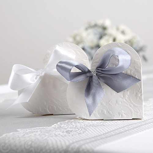 Dwa sercowe pudełka w kolorze białym, jedno z białą wstążką, a drugie z szarym kokardą, ozdobione delikatnym wypukłym wzorem, stojące na jasnym tle z kwiatami w tle.