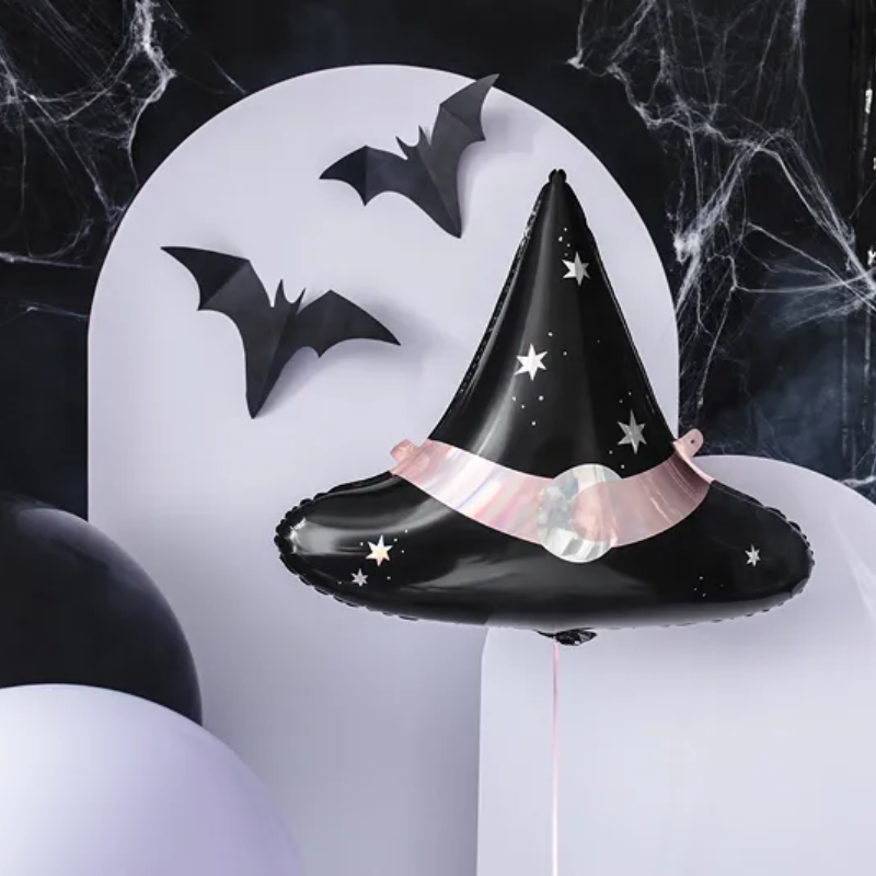 Czarny balon w kształcie czapki czarownicy z gwiazdkami, ozdobiony różową wstążką i okrągłym elementem, zawieszony na tle purpurowych i czarnych balonów oraz dekoracji w kształcie nietoperzy, z pajęczyną w tle.