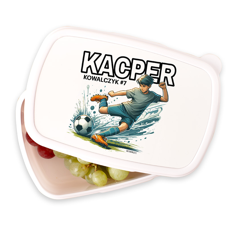 Lunch box with an illustration of a boy kicking a soccer ball, splashing water, and the text 'KACPER KOWALCZYK #7' on the lid; the box contains grapes and red fruits inside.