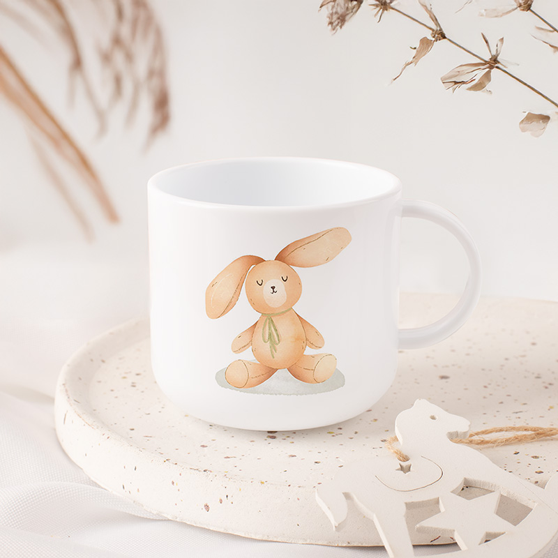 Biały kubek z ilustracją siedzącego królika w kolorze brązowym z dużymi uszami i zieloną kokardką, na jasnym tło z dekoracyjnymi elementami roślinnymi i ceramiczną podstawą.