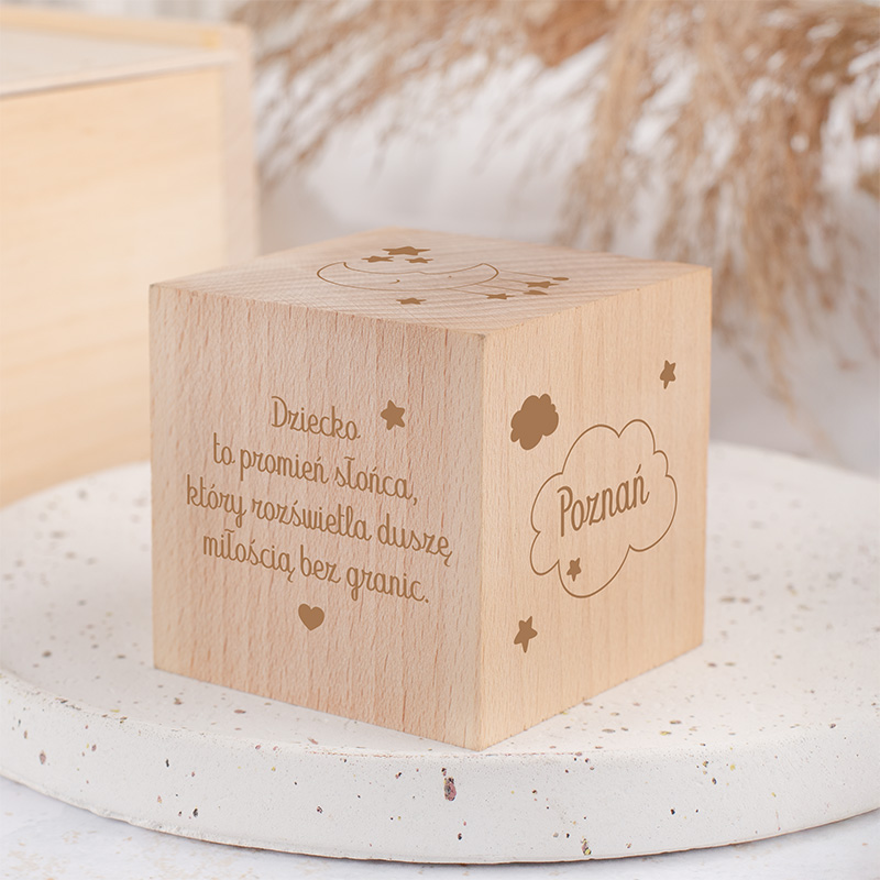 Drewniany sześcian z wygrawerowanym tekstem, w którym napisano: "Dziecko to promień słońca, który rozświetla duszę miłością bez granic" oraz chmurką z napisem "Poznań", otoczony przez gwiazdki. Sześcian znajduje się na jasnym, teksturowanym talerzu w tle z suszonymi roślinami.