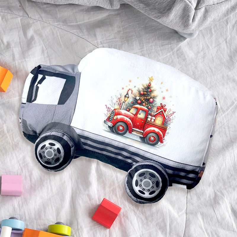 Poduszka w kształcie ciężarówki z nadrukiem czerwonego pickupa przewożącego choinkę i ozdoby świąteczne, leżąca na białej pościeli obok kolorowych klocków.