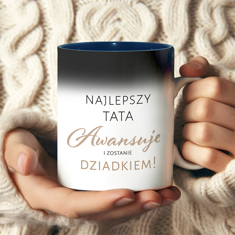 Kubek trzymany w rękach osoby w beżowym swetrze, z napisem 'Najlepszy Tata Awansuje i Zostanie Dziadkiem' na białym tle, z ciemnoniebieskim wnętrzem.