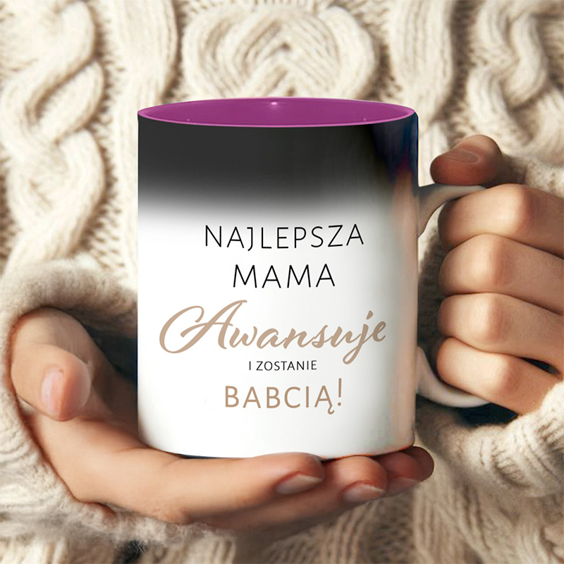 Kubek z napisem 'Najlepsza Mama Awansuje i Zostanie Babcią!' trzymany w dłoniach osoby w swetrze z podwiązanym wzorem na tle.