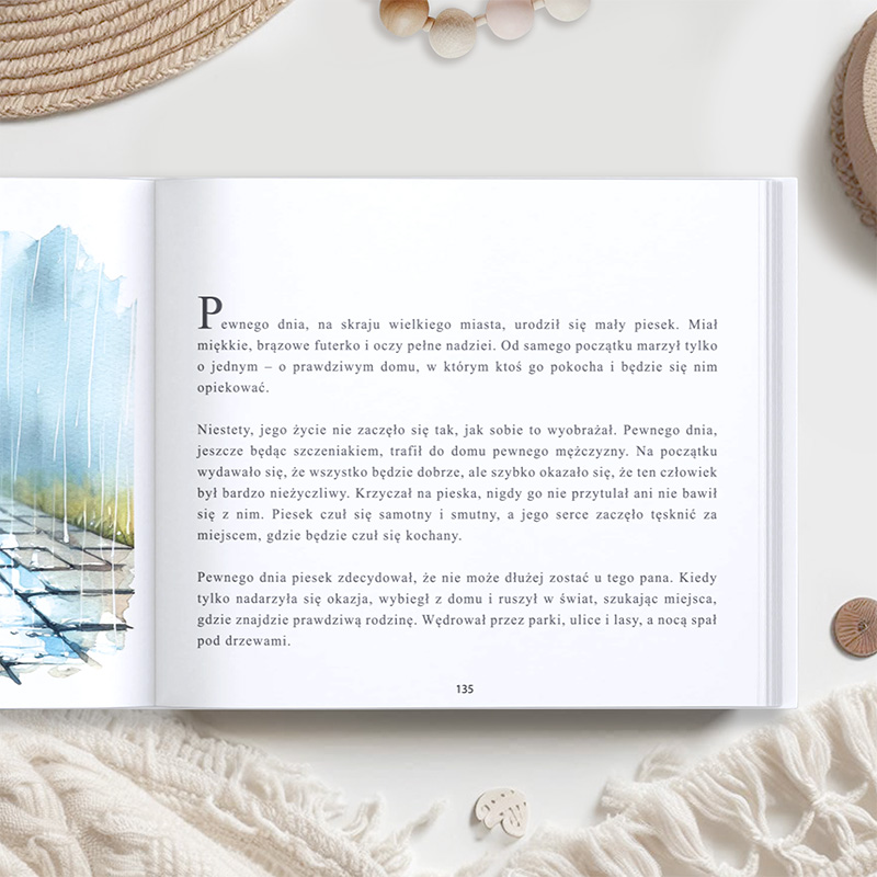 Otwarte książka z ilustrowaną stroną obok widoku na blejtram i kawałki sznurków dekoracyjnych. Ilustracja przedstawia delikatne niebieskie i brązowe akwarele, podczas gdy tekst dotyczy małego psa z nadzieją na prawdziwy dom.