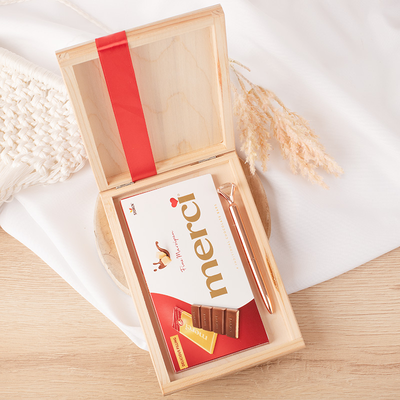 Otwarte drewniane pudełko z czerwonym paskiem, w którym znajduje się czekolada Merci, kawałki czekolady, rzemyk z napisem 'Merci' oraz różowa długopis; w tle delikatne rośliny i jasne tło.