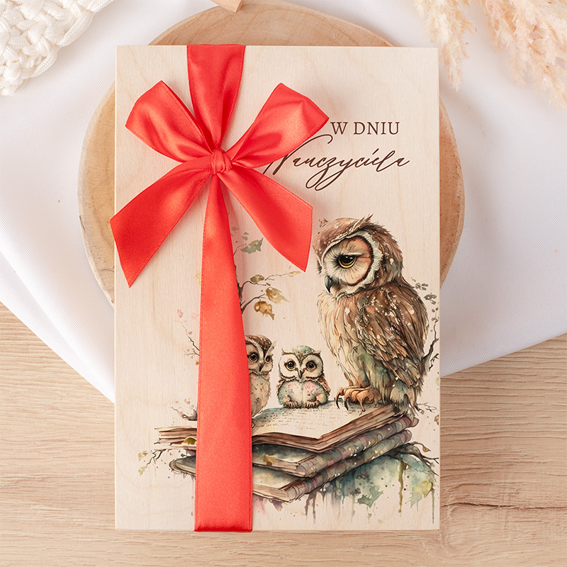 Elegancki notes z rysunkiem sów na tle książek, ozdobiony czerwoną wstążką, na drewnianej podstawie. Napisy na okładce w polskim języku.