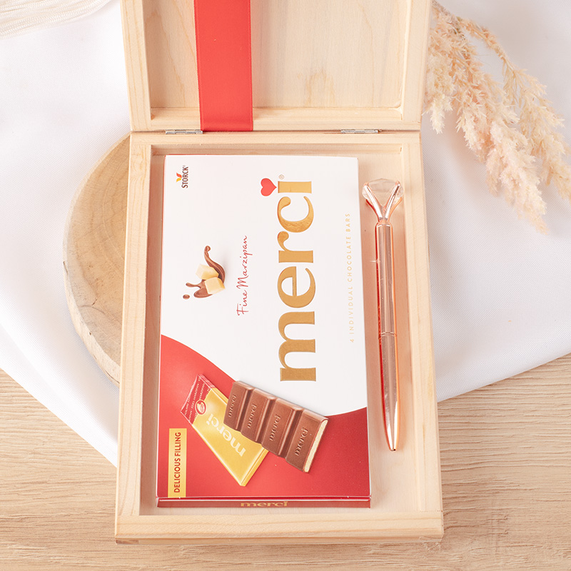 Otwarta drewniana skrzynka z zestawem czekoladek Merci, składającym się z opakowania z 4 indywidualnymi batonikami, obok znajduje się złoty długopis oraz mała biała karteczka z napisem Merci.
