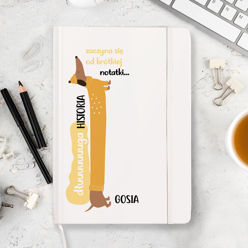 Notes z żółtą okładką z grafiką przedstawiającą psa rasy jamnik oraz napisem "dużunuga HISTORIA" i "zaczyna się od krótkiej notatki..." obok imienia "GOSIA", leżący na biurku obok długopisu i filiżanki kawy.