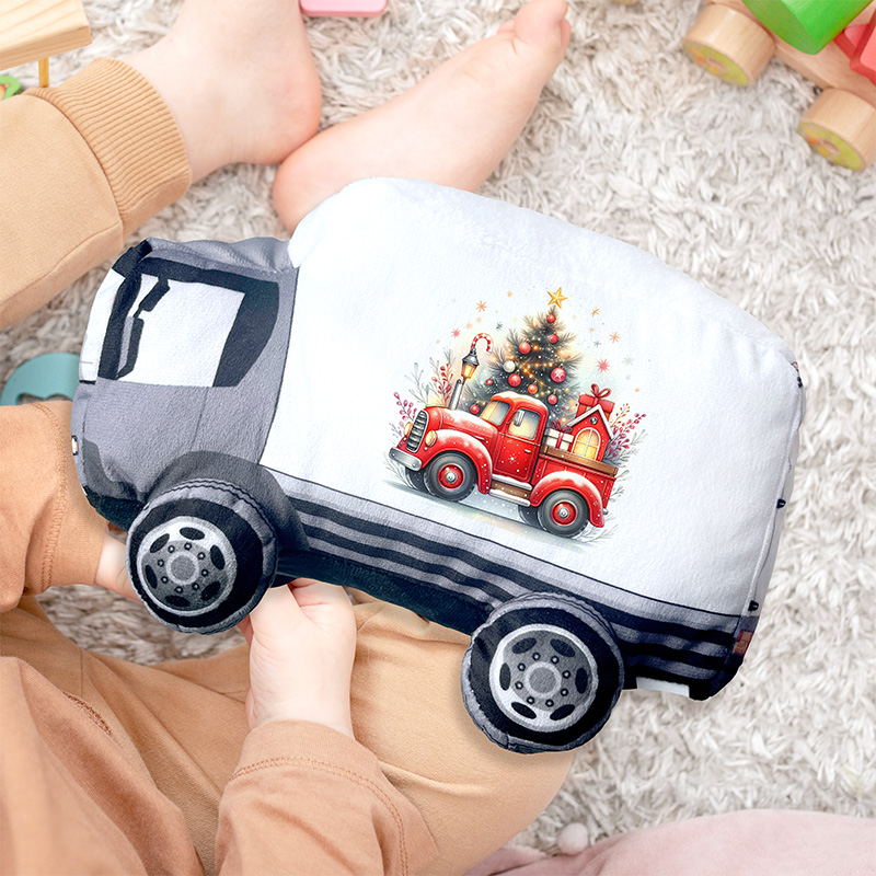Dwie ręce trzymają poduszkę w kształcie ciężarówki, na której znajduje się kolorowy nadruk z czerwoną ciężarówką ozdobioną choinką i prezentami, na tle puchatego dywanu z dziecinnymi zabawkami w pobliżu.