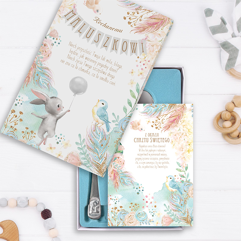 Zestaw prezentowy na chrzest z kolorowym opakowaniem, zawierający kartkę z ilustracją królika trzymającego balon oraz niebieskiego ptaka, na tle pastelowych wzorów i złotych akcentów. Wewnątrz widoczna łyżeczka i druga kartka z życzeniami z okazji chrztu.