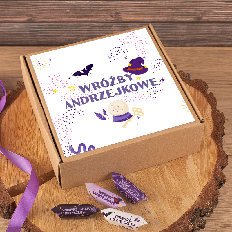 Zamknięte pudełko z napisem 'Wróżby Andrzejkowe' na pokrywie, ozdobione grafikami przedstawiającymi kapelusz czarownicy, nietoperza i klucz, leży na drewnianej podstawie z fioletową wstążką obok, a przy nim kilka kolorowych karteczek z wróżbami.