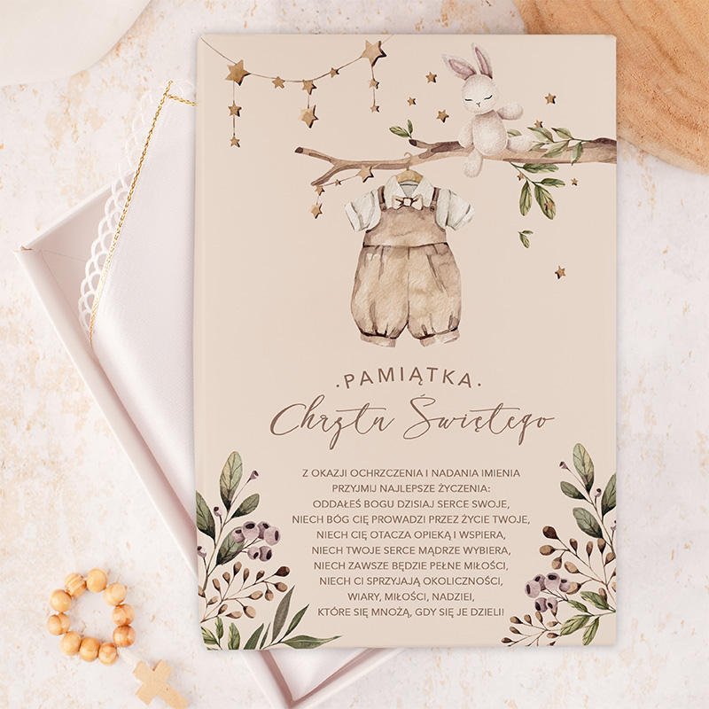 Kartka z okazji chrztu, z ilustracją ubranka dziecięcego zawieszonego na gałęzi, obok jasnobrązowego króliczka, z dekoracyjnymi gwiazdkami i liśćmi oraz napisem "Pamiątka Chrzest Świętego" w stylu kaligraficznym. Tekst życzeń na dole kartki.