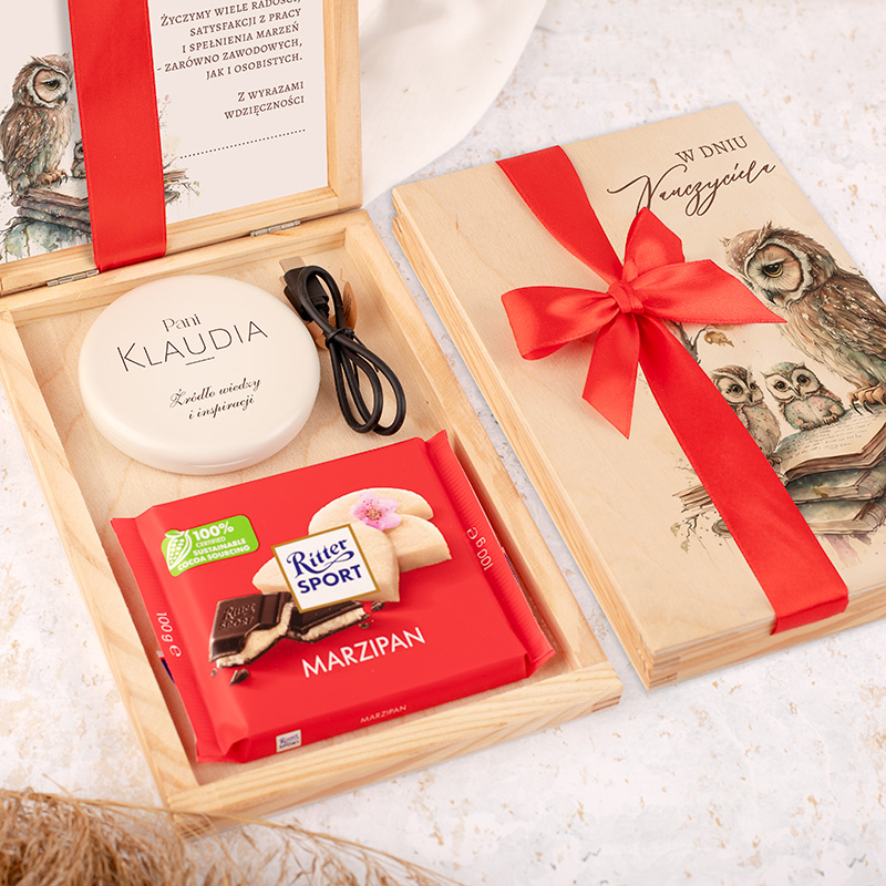 Elegancka drewniana skrzynka prezentowa z czerwonym kokardą, zawierająca krem do ciała z imieniem "Pani Klaudia", kabel oraz paczkę czekolady Ritter Sport z marcepanem.