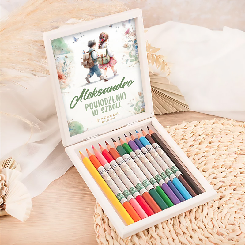 Biały drewniany pojemnik z kredkami w różnych kolorach, z otwartą wieczkiem, na którym znajduje się ilustracja dzieci idących w stronę szkoły z napisem "Aleksandro, powodzenia w szkole". Tło stanowi jasna, pleciona podkładka.