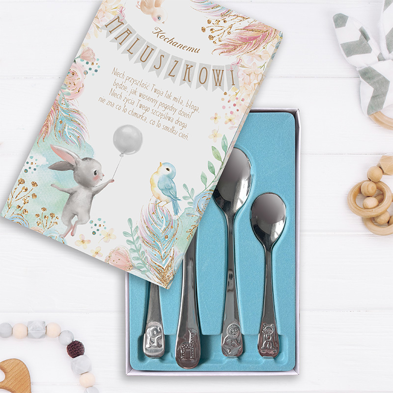 Zestaw czterech srebrnych łyżeczek w pudełku z niebieskim wyściółką, obok kolorowa kartka z napisem 'Kochanemu Maluszkowi' oraz ilustracjami zwierząt; w tle są drewniane zabawki i dekoracyjne koraliki.