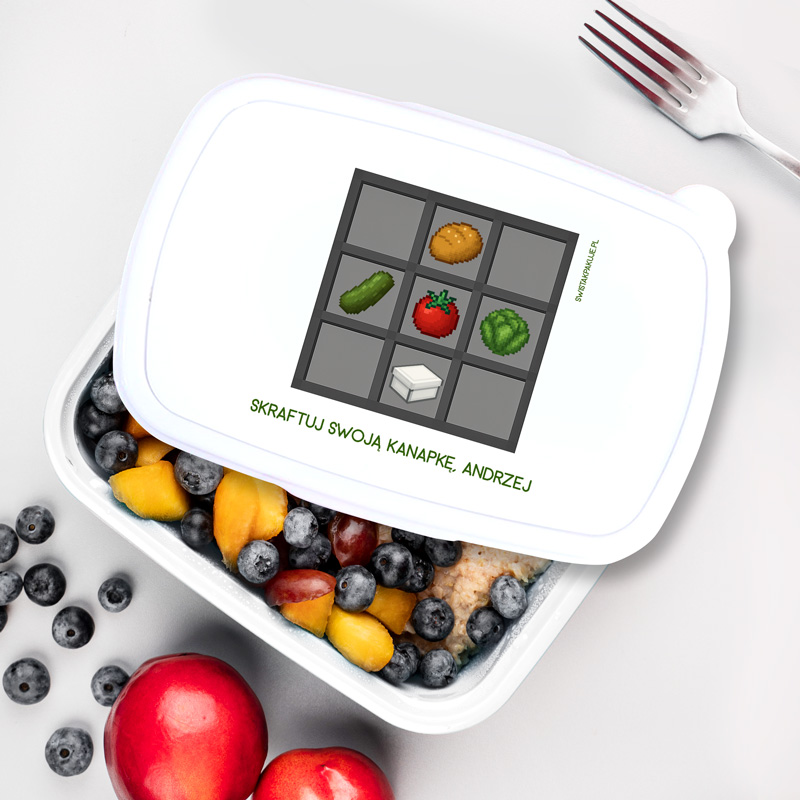 Pojemnik na jedzenie z ilustracją przedstawiającą siatkę do tworzenia kanapek, zawierającą różne składniki takie jak ogórek, pomidor i sałata, z napisem 'SKRAFTUJ SWOJA KANAPKĘ, ANDRZEJ' na pokrywce, otoczony kolorowymi owocami i widelcem obok.