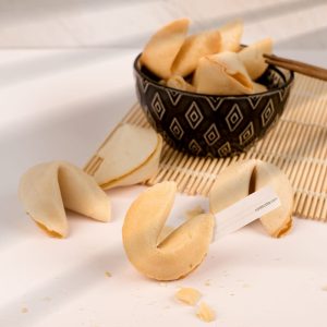 Fortune cookies scattered on a light surface, with a dark bowl containing more cookies in the background and a bamboo mat underneath.