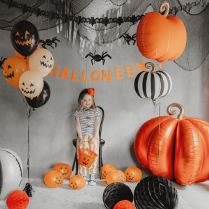 Dziecko w pasiastych spodniach stoi na krześle w otoczeniu balonów na Halloween, w tym balonów w kształcie dyni oraz dużej, pomarańczowej, nadmuchanej dyni. Na tle znajduje się dekoracja z pajęczyną i napis 'HALLOWEEN' w pomarańczowym kolorze.