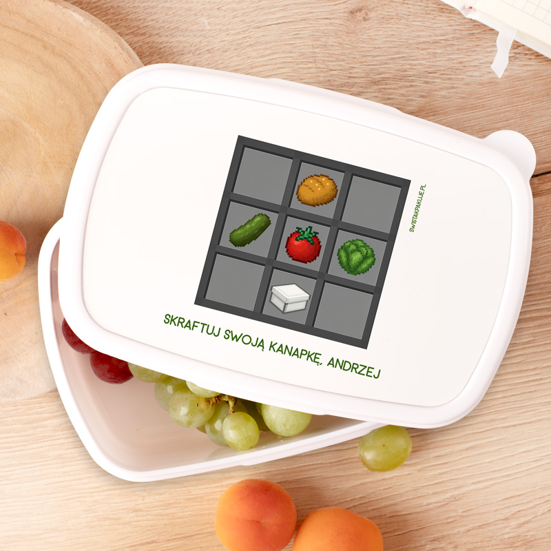 Białe pudełko na jedzenie z grafiką przedstawiającą siatkę z różnymi składnikami: ogórkiem, pomidorem, sałatą, ziemniakiem oraz serem, napis "SKRAFTUJ SWOJA KANAPKE, ANDRZEJ" w zielonym kolorze, leżące na drewnianym blacie obok owoców.