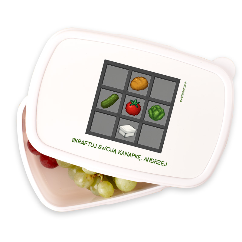 Pojemnik na jedzenie z grafiką przypominającą interfejs gry, przedstawiający składniki do kanapki: ogórek, pomidor, sałata, bułka i ser, z napisem 'SKRAFTUJ SWOJA KANAPKE, ANDRZEJ' w dolnej części. Wewnątrz pojemnika widoczne zielone i czerwone winogrona.