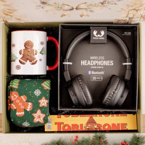 Zestaw upominkowy zawierający kubek z wzorem piernikowego człowieka, bezprzewodowe słuchawki oraz ciepłe skarpetki z piernikami, ułożony na tle pudełka z czekoladą Toblerone.