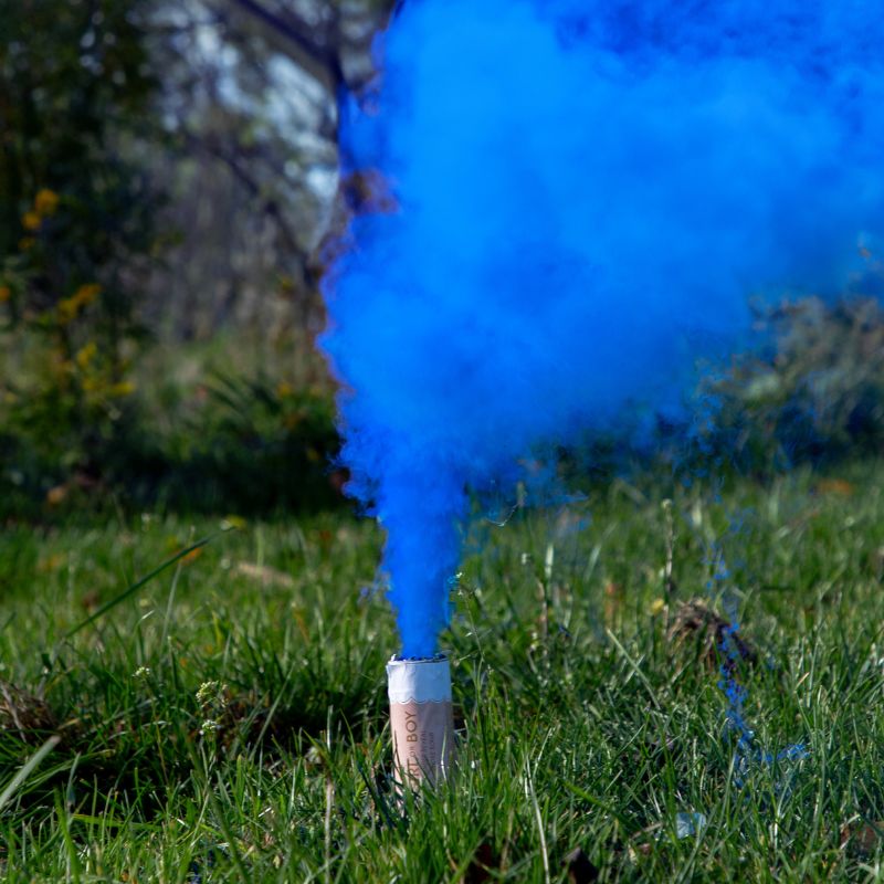 Fajerwerk z niebieskim dymem unoszącym się z pojemnika stojącego na zielonej trawie, w tle widać naturę i drzewka.