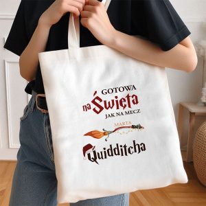 Kobieta trzyma białą torbę bawełnianą, na której znajdują się napisy: 'GOTOWA na Święta jak na mecz', imię 'MARTA' oraz słowo 'Quidditcha', obok którego jest graficzna ilustracja miotły i elementy świąteczne.
