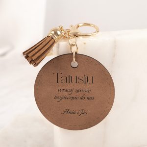 Brązowy, okrągły breloczek z napisem 'Tatusiu', z dodatkowymi słowami 'wracaj zawsze bezpiecznie do nas' oraz podpisem 'Ania i Jas', zakończony frędzelkiem.
