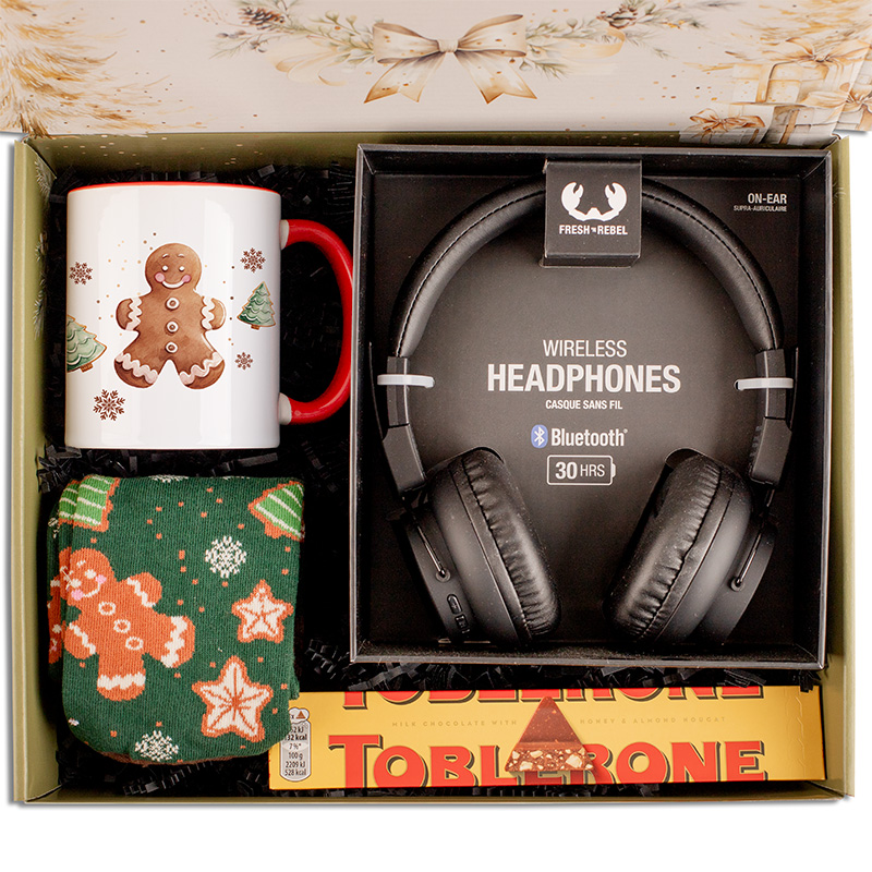 Zestaw prezentowy zawierający kubek z wizerunkiem piernika, bezprzewodowe słuchawki oraz zielone skarpetki z motywem piernika, obok czekoladki Toblerone na dnie pudełka.