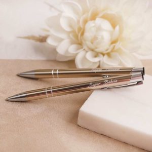 Dwa metalowe długopisy w złotym kolorze leżą na jasnym tle, obok znajduje się biały kawałek mydła i jasna kwiatowa dekoracja.