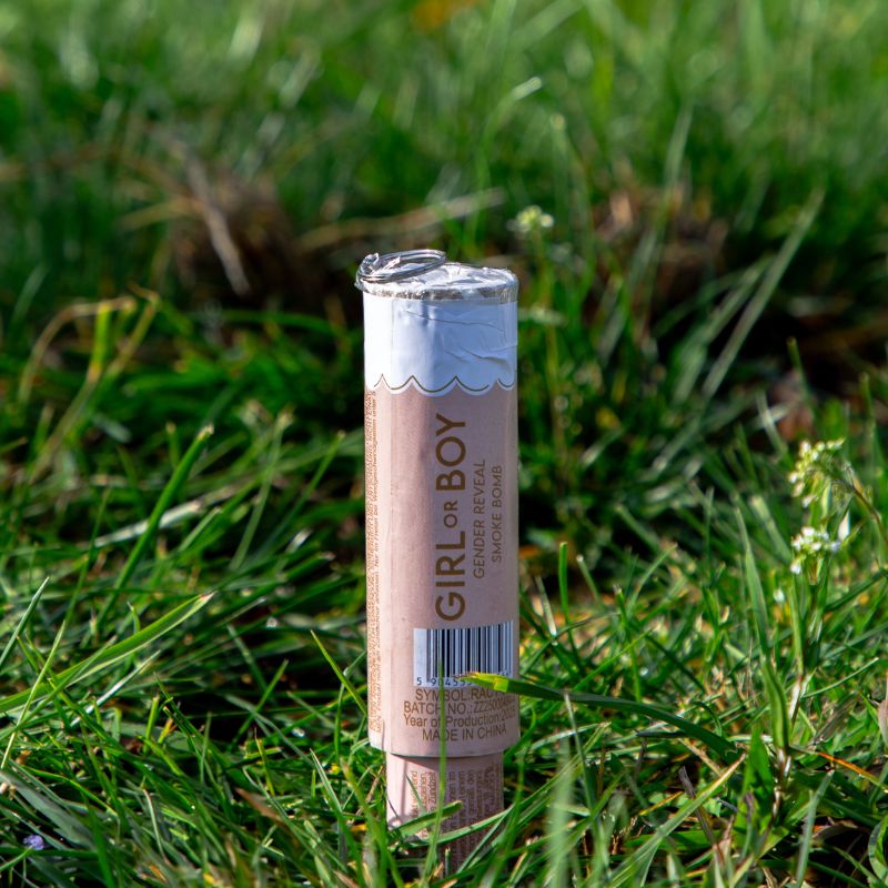 Rurka z napisem 'GIRL or BOY' pełniąca funkcję bombki dymnej do ujawnienia płci, leżąca na trawie.