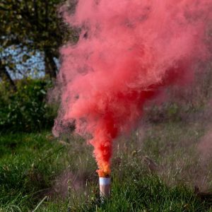 Czerwony dym wydobywający się z tuby w trawie, otoczony zielonymi krzakami i drzewami w tle.