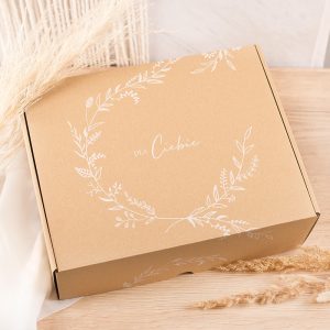 Brązowy kartonowy pudełko ozdobione białymi elementami roślinnymi i napisem "DLA Ciebie" na górze, umieszczone na jasnym drewnianym blacie obok suchych traw.