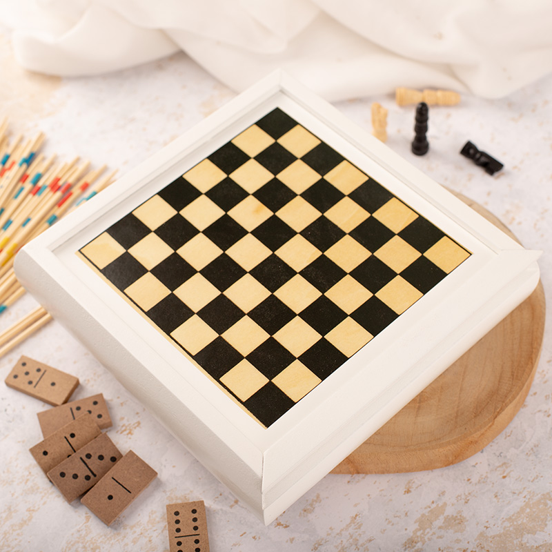 Zbliżenie planszy do gry w szachy w białej ramie, umieszczonej na drewnianym podkładzie, otoczonej kostkami domina i zapałkami.