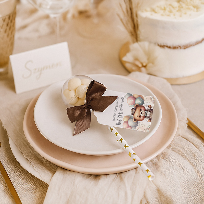 Ozdobne miejsce na stole z białym talerzem i różowym talerzem podkładowym, na którym znajduje się przeźroczysta kulka z białymi słodyczami, przewiązana brązową wstążką; obok leży papierowa łyżka z grafiką. W tle widoczny tort oraz elegancka karta z imieniem 'Szymon'.