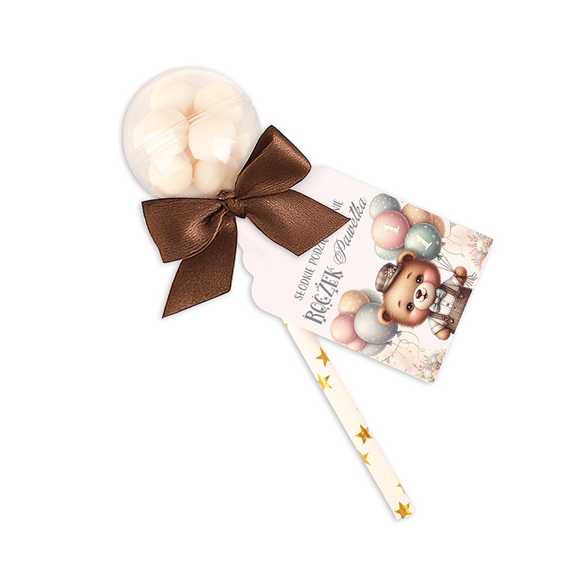 Słodka niespodzianka w formie lizaka składającego się z przezroczystej kuli zawierającej białe słodycze, przyozdobionego brązową wstążką i kartką z ilustracją misia trzymającego balony oraz napisem.